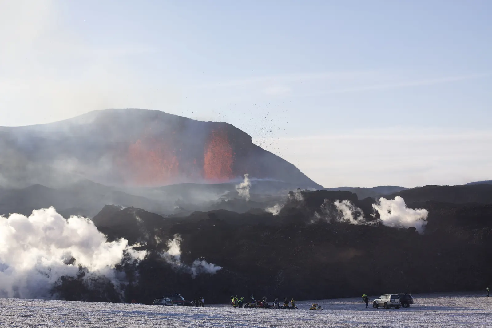A fountain of red lava rising from a black ridge, with white steam clouds below and people on the snow in front
