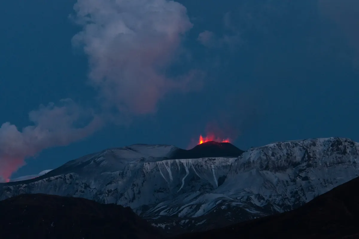 A small crater glowing red at dusk on a snow-covered mountain pass, with a grey plume of steam rising beside it