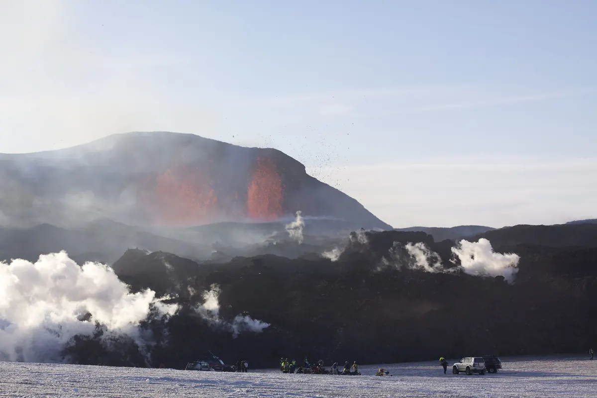 A fountain of red lava rising from a black ridge, with white steam clouds below and a few people standing on the snow in front