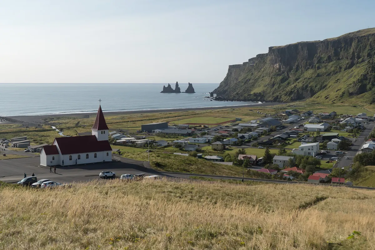 The white church with a red roof above the village of Vík, with a black sand beach and three sea stacks behind it