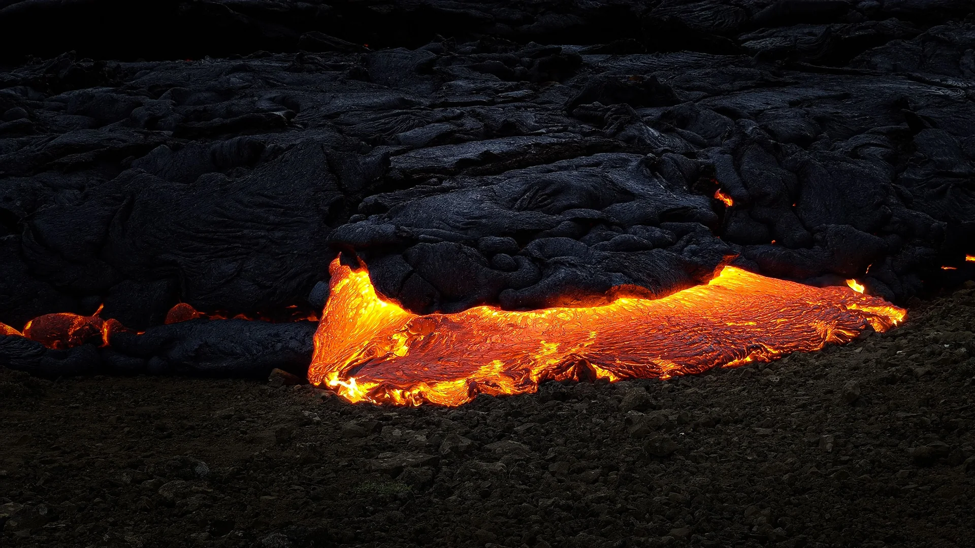 Glowing orange lava flowing out from under a crust of black rock on an Icelandic lava field