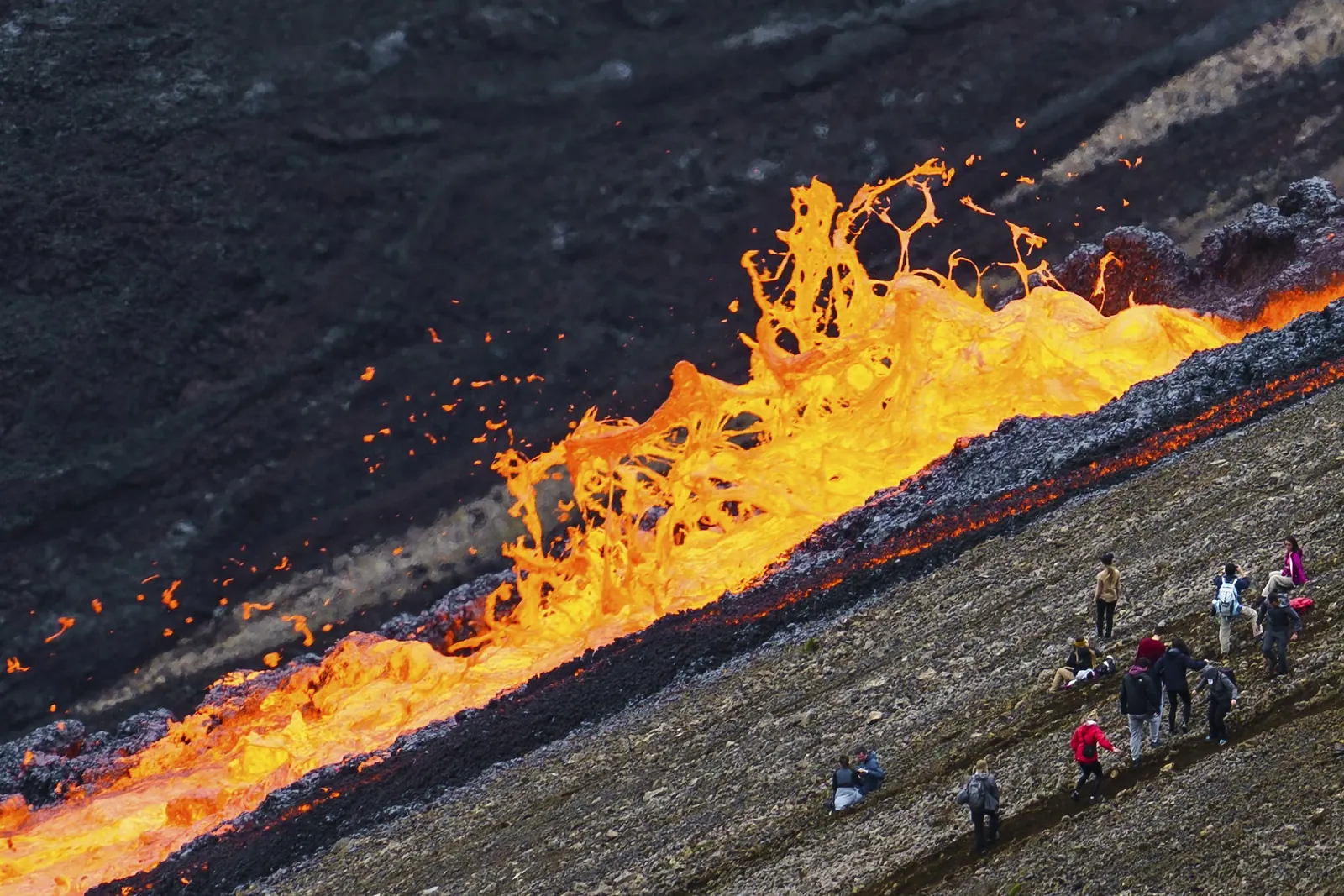 A wave of bright orange lava surges along a ridge while hikers watch from the slope below