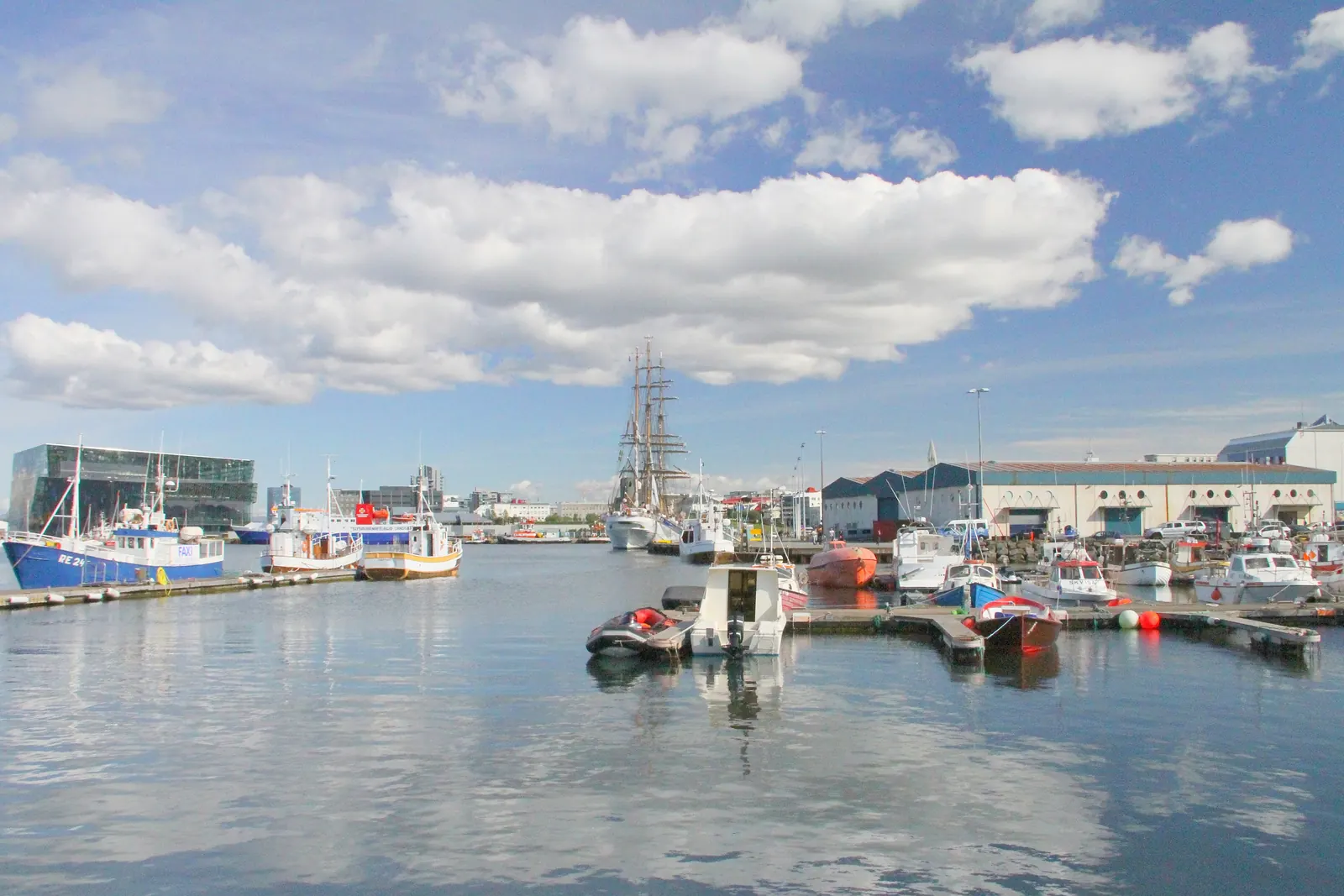 Fishing boats and a tall ship moored in Reykjavík's Old Harbour on a bright day, with the glass facade of Harpa at the left