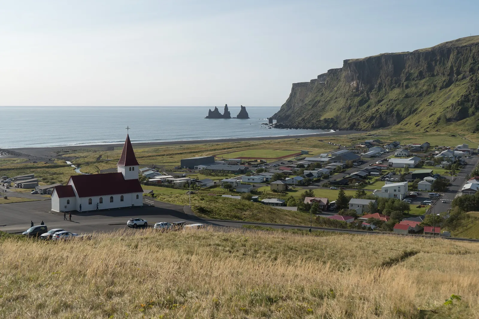 The white church with a red roof above the village of Vík, with a black sand beach and three sea stacks behind it